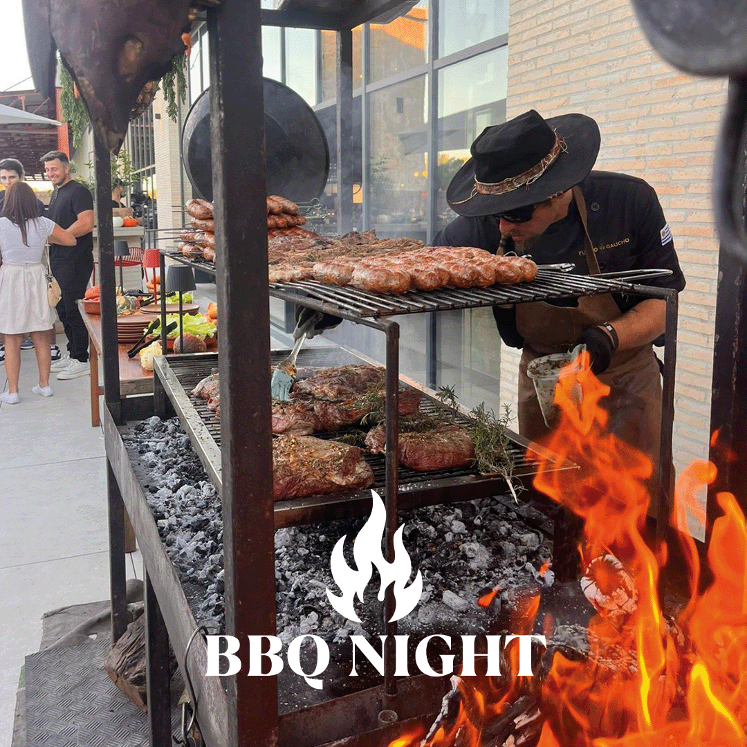 Maestro de barbacoa con sombrero negro y equipo de cervecero da la vuelta a la carne asada sobre un fuego abierto, junto a una barbacoa con trozos de carne dorados, llamas e invitados al fondo.